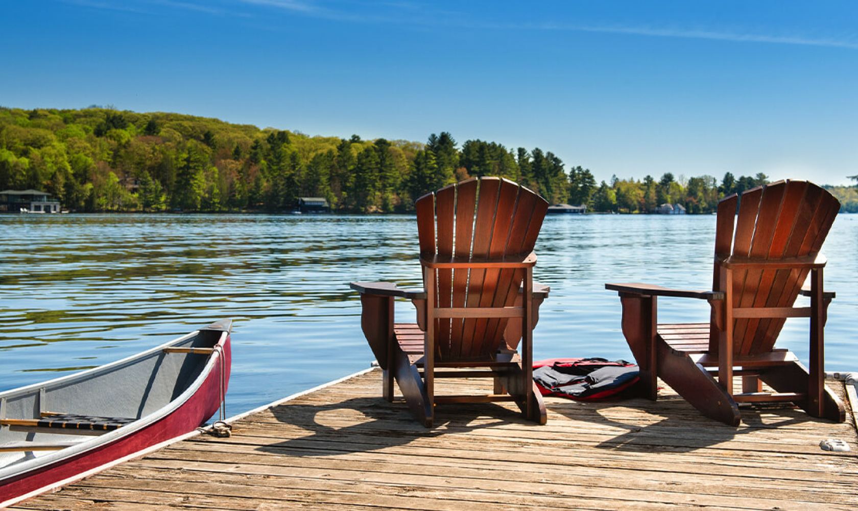 Two wooden chairs sit on a lakeside dock, next to a canoe.