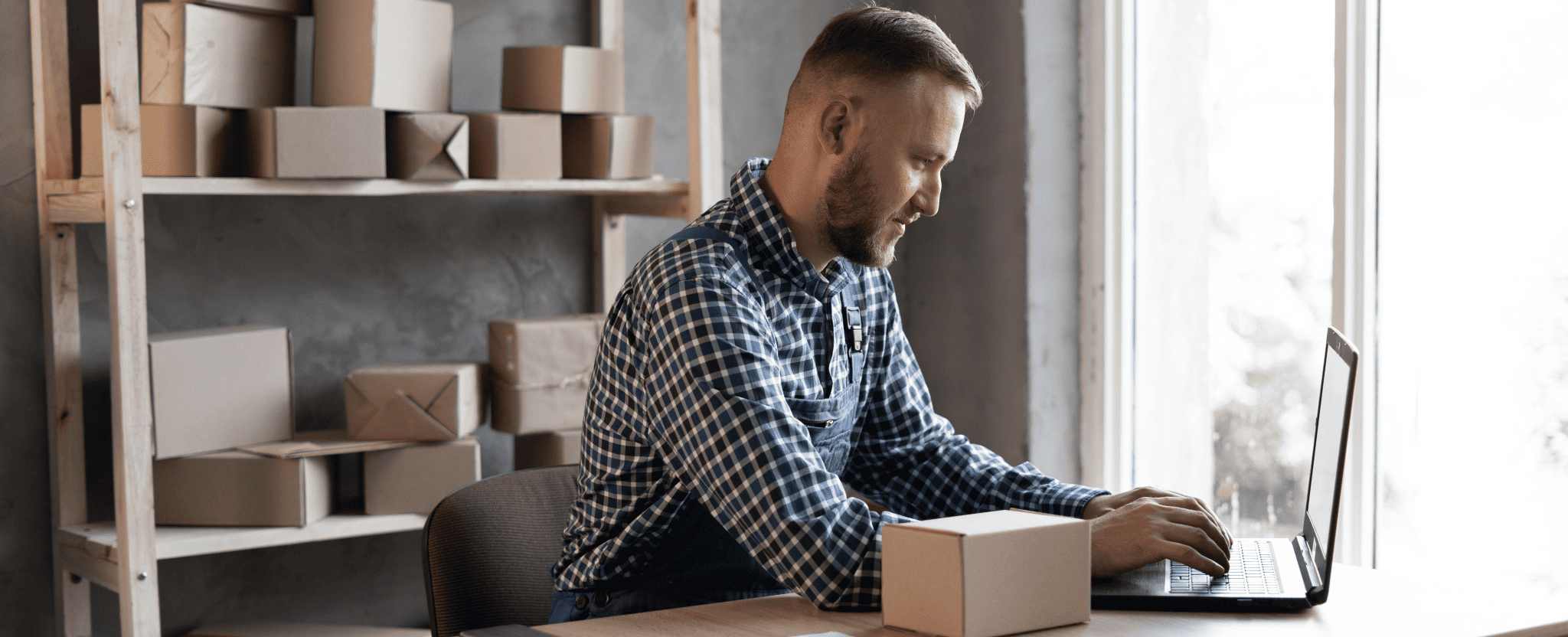 A man sits at a desk and types on a laptop in a room full of boxes.