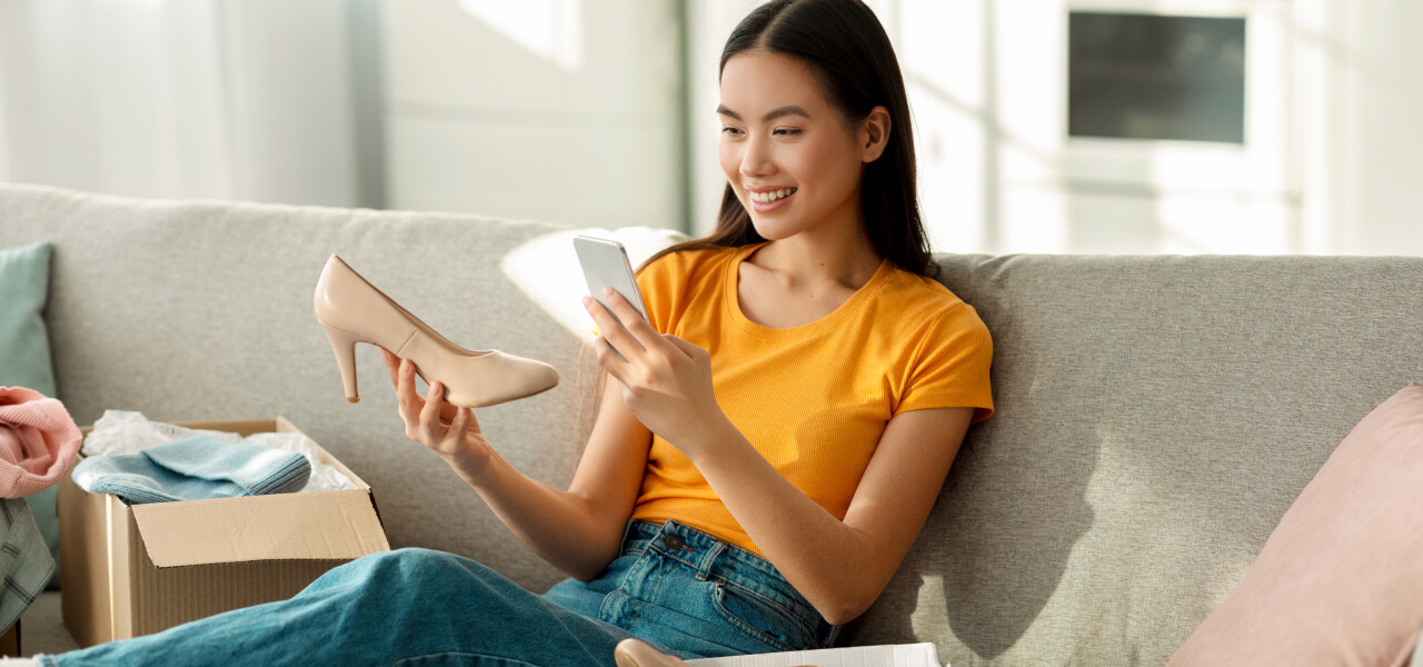 A smiling young woman sits on a sofa, holding a new shoe in one hand and a smartphone in the other. There’s an open shipping box next to her.