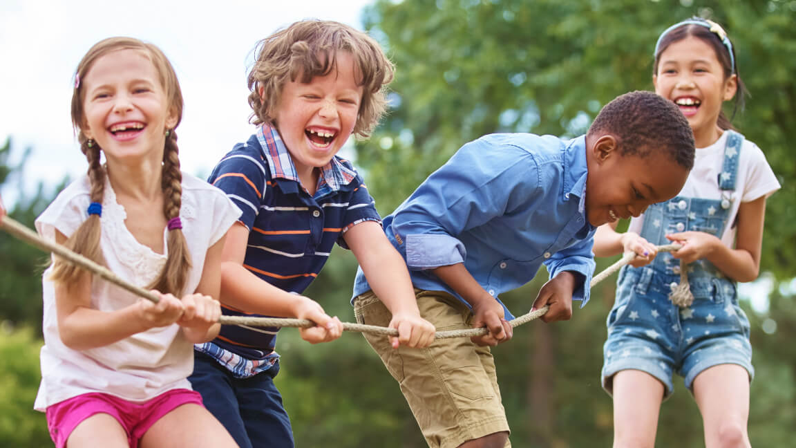 Four children in a park laugh as they hold on to a tug-of-war rope.