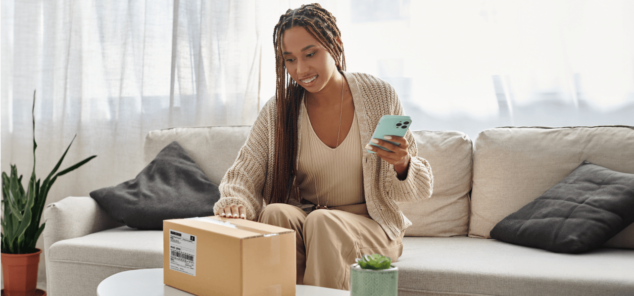 Une femme souriante regarde son téléphone intelligent en tenant un colis.