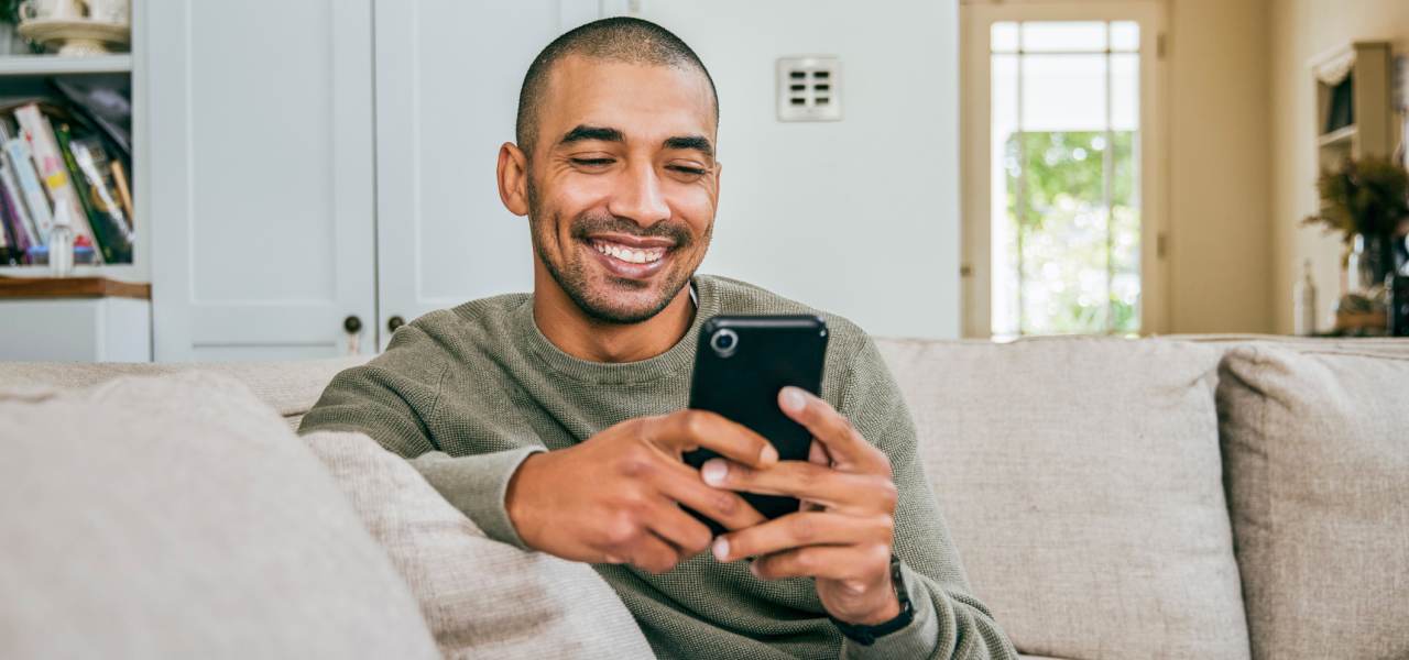 Un jeune homme sourit et fixe un téléphone cellulaire qui se trouve dans ses mains. Il est assis sur un canapé dans son salon.