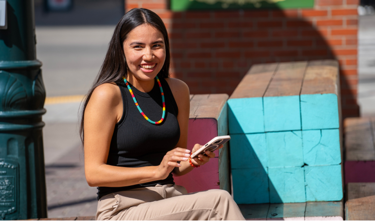 Une femme assise sur un banc dans un espace urbain extérieur, tenant son téléphone intelligent et souriant.