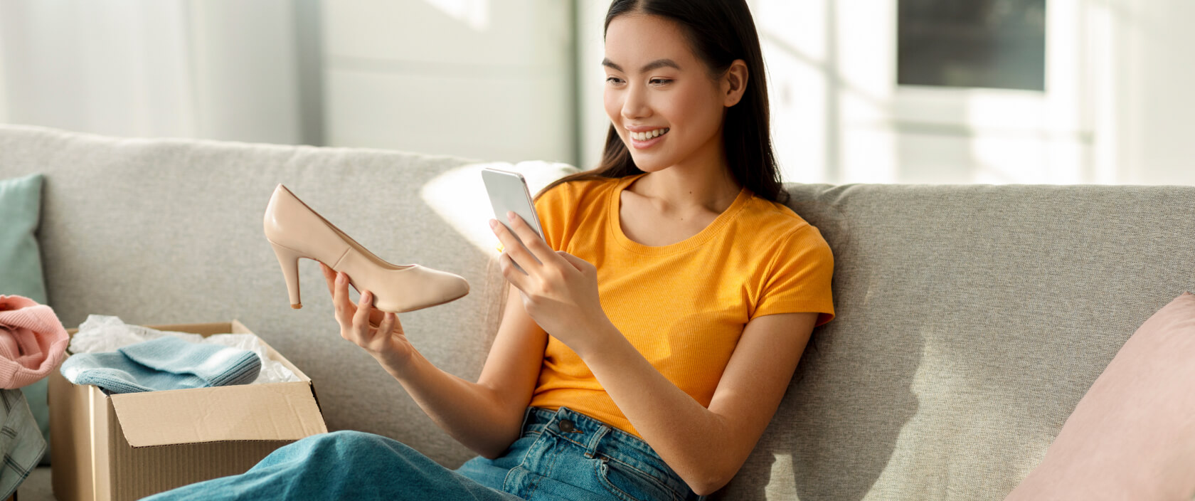 Assise sur son sofa, une jeune femme tout sourire photographie une nouvelle chaussure avec son téléphone.