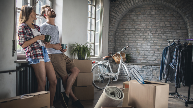 Un homme et une femme s’appuient contre un rebord de fenêtre dans un appartement rempli de boîtes de déménagement. 