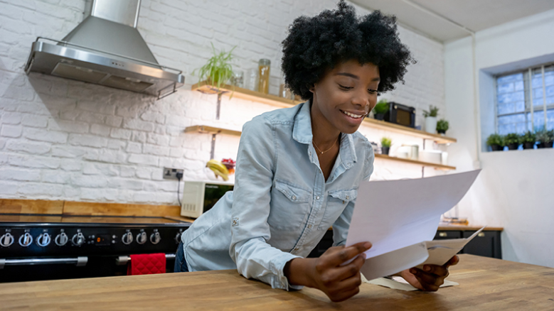 Une femme penchée contre son comptoir de cuisine sourit en lisant son courrier.