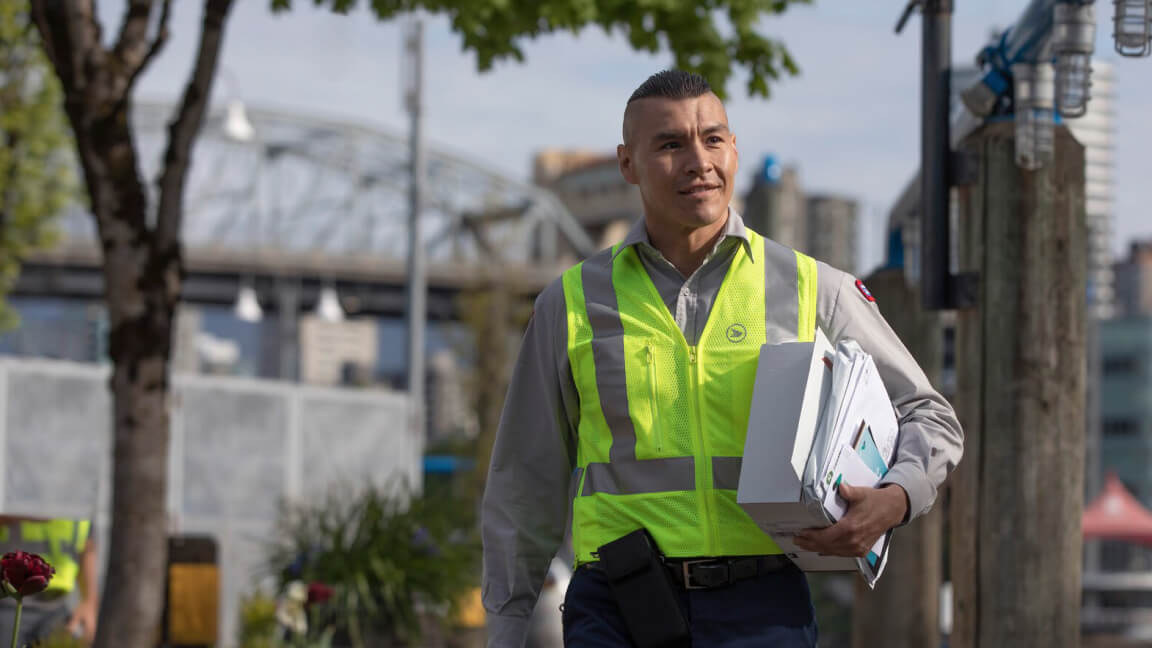 Un employé de Postes Canada vêtu d’un gilet de sécurité transporte du courrier. Il y a un pont et une ville en arrière-plan. 