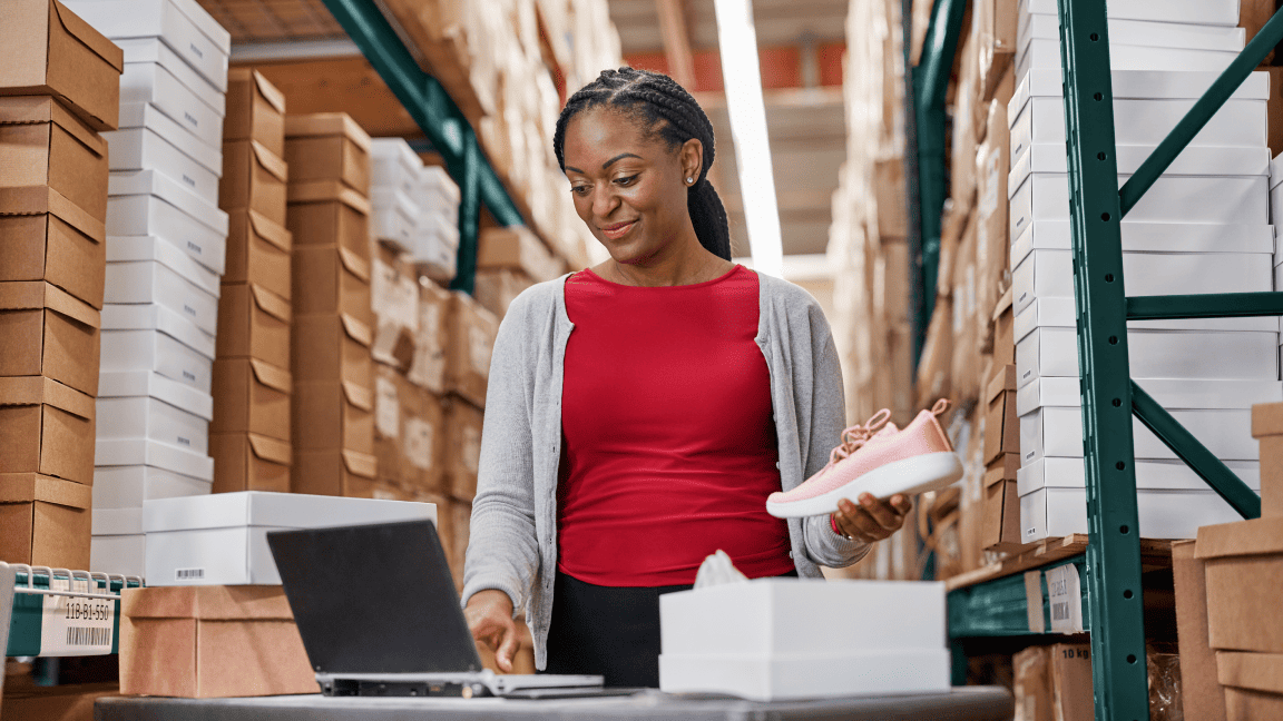 Une femme tape sur le clavier d’un ordinateur portable dans un grand entrepôt rempli de hautes piles de boîtes.